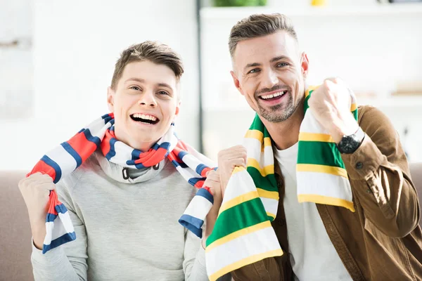 Retrato de sorrindo pai e filho adolescente em scarfs assistindo jogo de esporte em casa — Fotografia de Stock