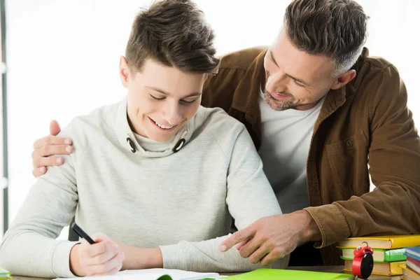 Sonriente padre abrazando alegre adolescente hijo mientras él haciendo tarea - foto de stock