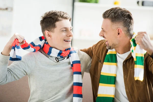 Happy father and teen son in scarfs watching sport match at home, looking at each other — Stock Photo