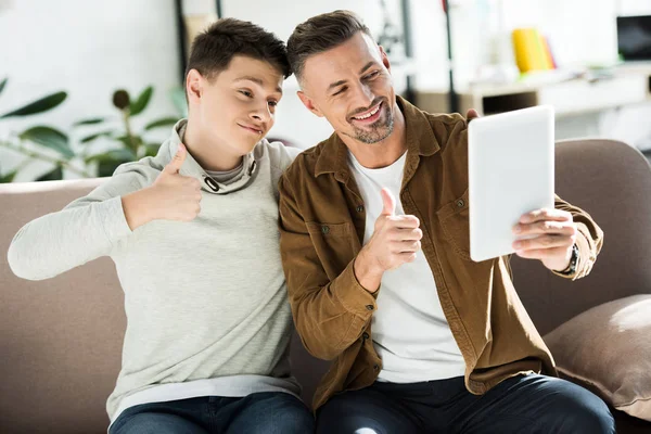 Lächelnder Vater und Teenager-Sohn schauen auf Tablet und zeigen zu Hause Daumen hoch — Stockfoto