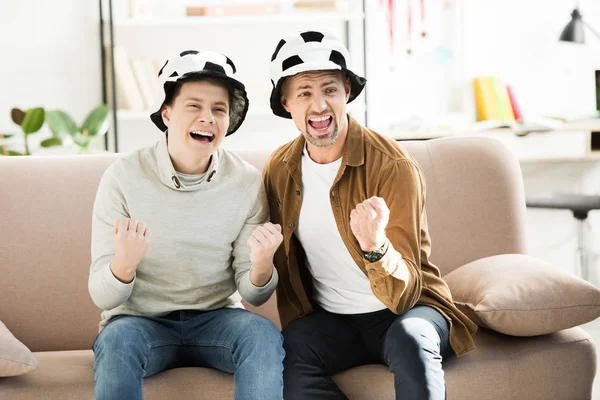 Excité père et adolescent fils dans le football chapeaux regarder jeu et crier sur canapé à la maison — Photo de stock