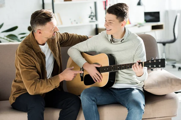 Sonriente hijo adolescente tocando la guitarra acústica para el padre en casa - foto de stock