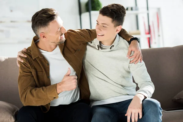 Sonriente padre e hijo adolescente abrazándose en el sofá en casa y mirándose — Stock Photo