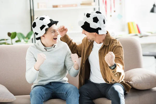 Excited father and teen son in football hats watching game and screaming on sofa at home, looking at each other — Stock Photo