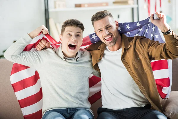Збуджений батько і підліток син, загорнутий в єдині штати прапор кричить на дивані під час спортивного матчу — стокове фото