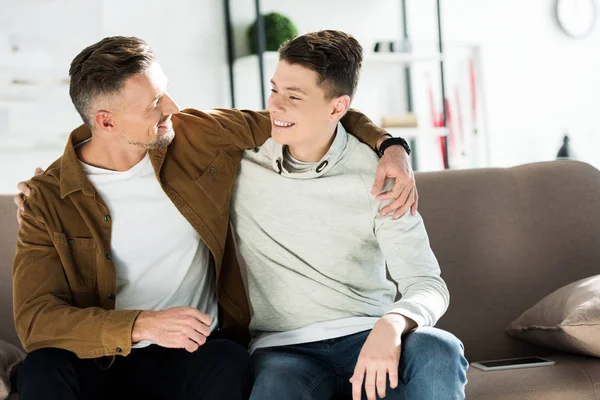 Sonriente padre e hijo adolescente abrazándose en el sofá en casa y mirándose - foto de stock