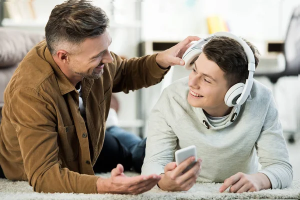 Smiling teen son lying on floor and showing something on smartphone to father — Stock Photo