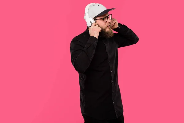 Elegante barbudo hipster masculino con anteojos y sombrero gorra escuchar música en auriculares aislados en rosa - foto de stock