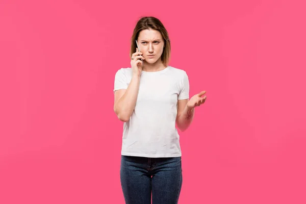 Irritated young woman talking on smartphone and gesturing by hand isolated on pink — Stock Photo