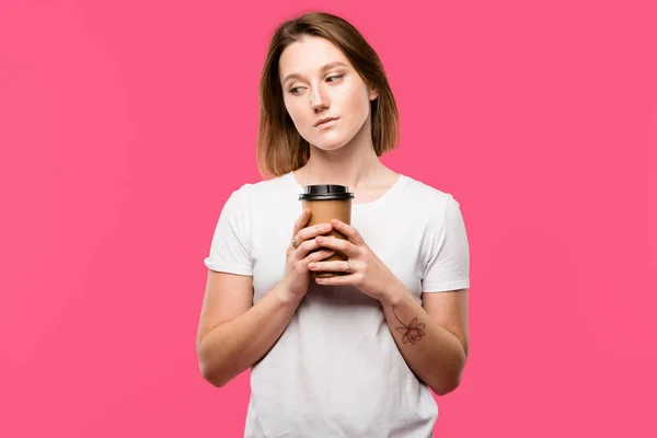 Задумчивая молодая женщина держит одноразовую чашку кофе изолированы на розовый — стоковое фото