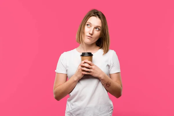 Мрійлива дівчина тримає одноразову чашку кави ізольовано на рожевому — стокове фото