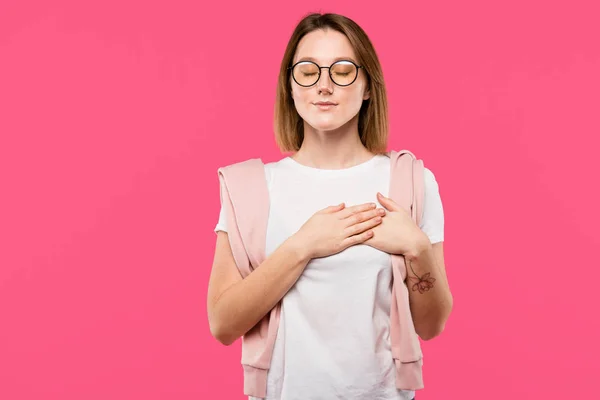 Мрійлива дівчина в окулярах тримає руки на грудях ізольовані на рожевому — стокове фото