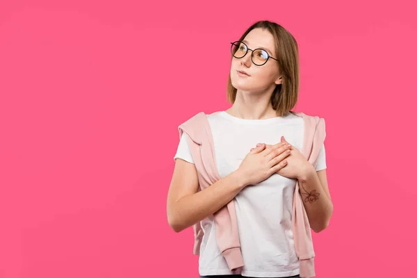 Fille rêveuse dans des lunettes tenant la main sur la poitrine et regardant loin isolé sur rose — Photo de stock