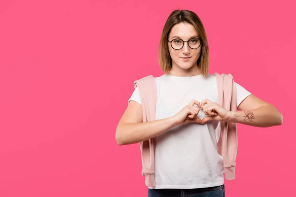 Fille élégante dans des lunettes montrant symbole de coeur isolé sur rose — Photo de stock