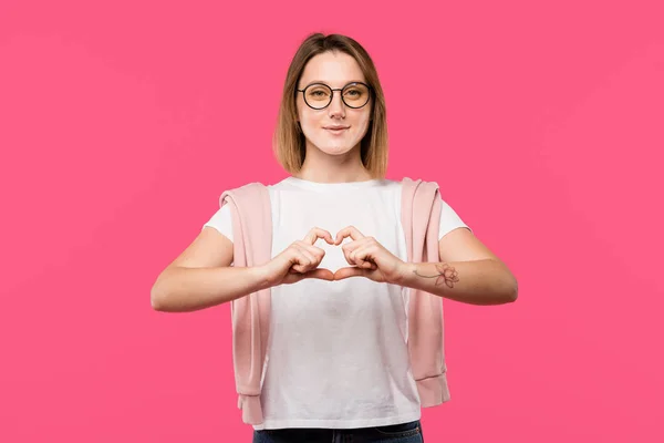 Sonriente chica con estilo en gafas que muestra el símbolo del corazón aislado en rosa - foto de stock