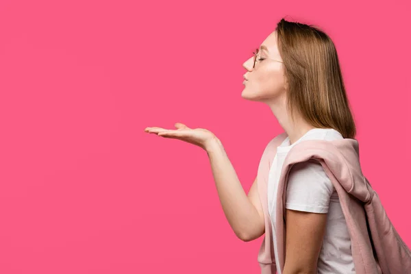 Side view of young woman with closed eyes blowing air kiss isolated on pink — Stock Photo