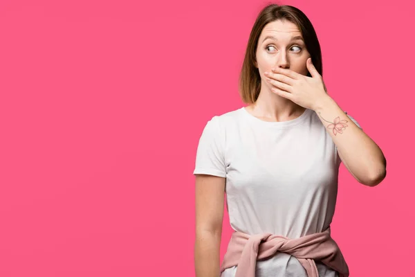 Sorprendida joven mujer tatuada cubriendo boca a mano aislada en rosa - foto de stock