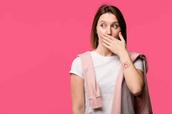 Schockierte junge tätowierte Frau bedeckt Mund von Hand isoliert auf rosa — Stockfoto