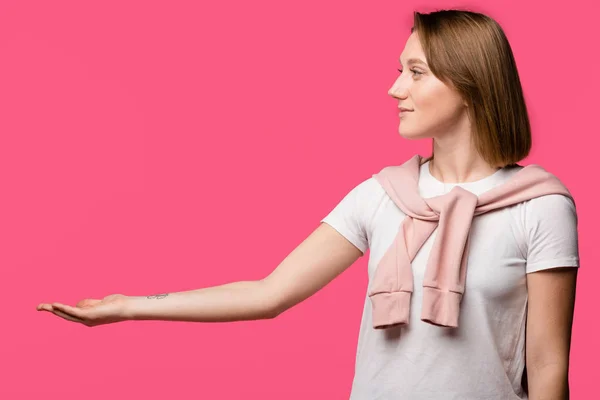 Вид збоку усміхнена дівчина тримає відкриту руку долоні ізольовано на рожевому — стокове фото