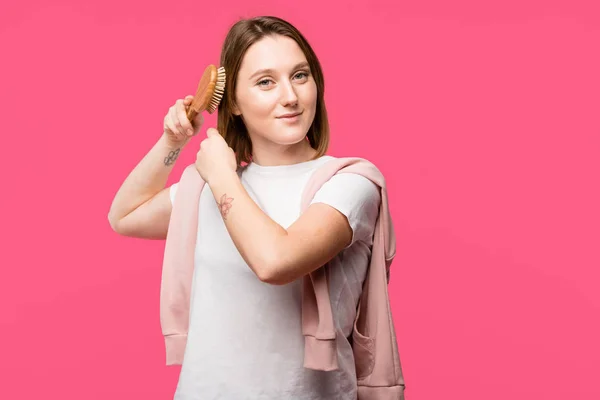 Cheerful beautiful girl brushing hair and looking at camera isolated on pink — Stock Photo