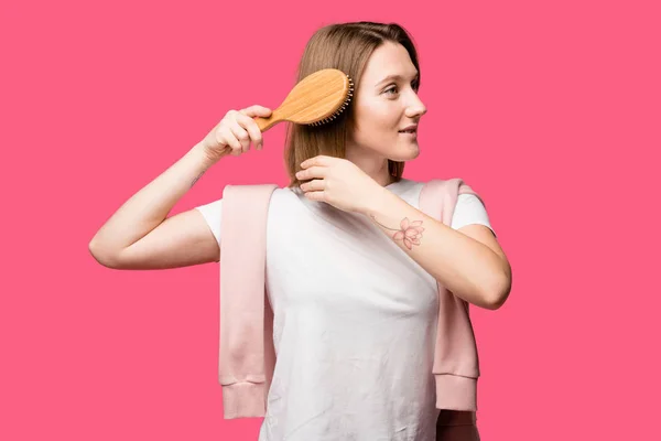 Sorridente jovem penteando cabelo e olhando para longe isolado em rosa — Fotografia de Stock