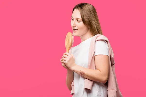 Bela jovem segurando escova de cabelo e cantando isolado em rosa — Fotografia de Stock