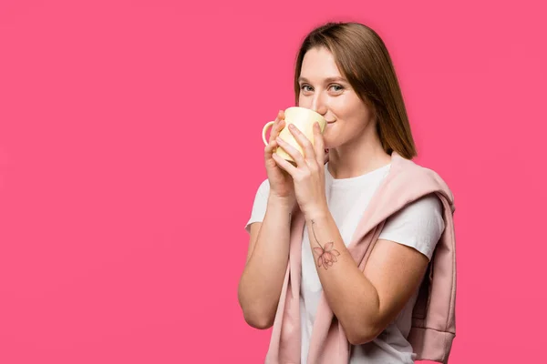 Giovane donna che beve dalla tazza e guarda la fotocamera isolata sul rosa — Foto stock