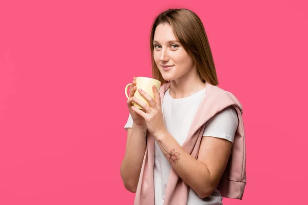 Jeune femme tenant tasse et souriant à la caméra isolé sur rose — Photo de stock