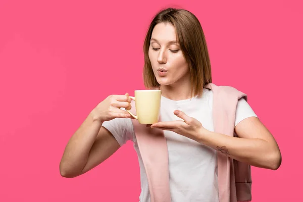 Молода жінка тримає чашку і дме на гарячому напої ізольовано на рожевому — стокове фото