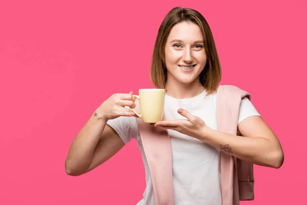 Attrayant jeune femme tenant tasse et souriant à la caméra isolé sur rose — Photo de stock