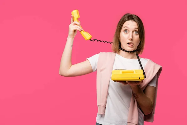 Mujer joven con alambre alrededor del cuello sosteniendo el teléfono giratorio y mirando a la cámara aislada en rosa - foto de stock