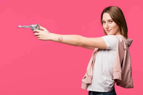 Vue latérale de la jeune femme tenant revolver et souriant à la caméra isolée sur rose — Photo de stock