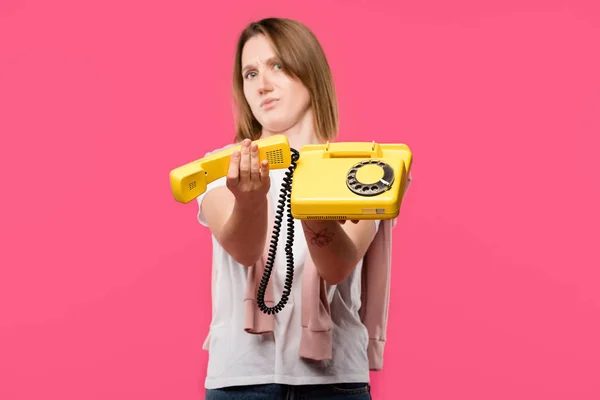 Молода жінка з кидаючим обличчям тримає жовтий роторний телефон ізольовано на рожевому — стокове фото