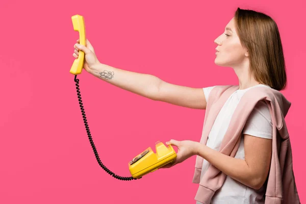 Вид сбоку на молодую женщину с желтым вращающимся телефоном, изолированным на розовом — стоковое фото
