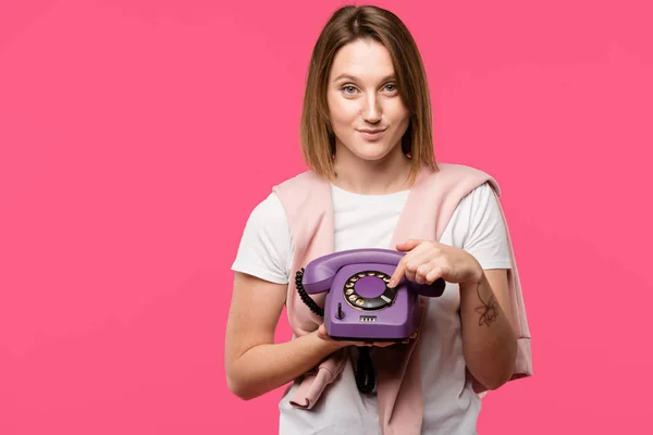 Beautiful young woman holding rotary phone and smiling at camera isolated on pink — Stock Photo