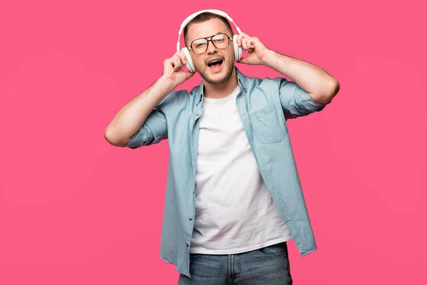 Happy young man in eyeglasses listening music in headphones and smiling at camera isolated on pink — Stock Photo