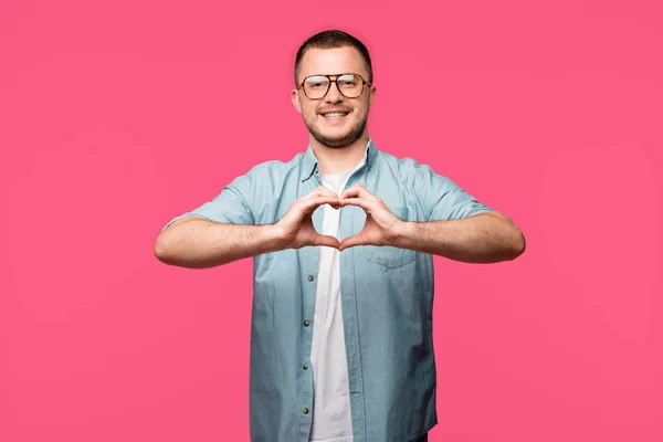 Happy man in eyeglasses showing hand heart symbol and smiling at camera isolated on pink — Stock Photo