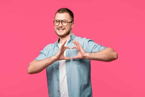 Счастливый молодой человек показывая руку символ сердца и улыбаясь в камеру изолированы на розовый — стоковое фото