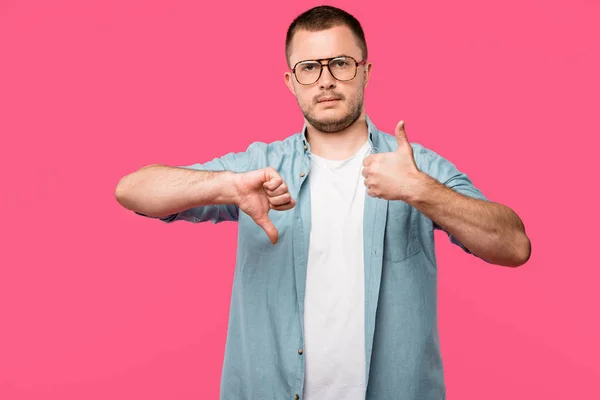 Uncertain man in eyeglasses showing thumb up and thumb down isolated on pink — Stock Photo