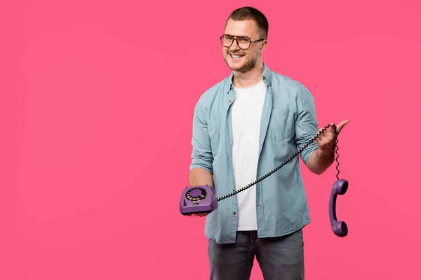 Joven guapo en gafas con el teléfono giratorio púrpura y sonriendo a la cámara aislado en rosa - foto de stock