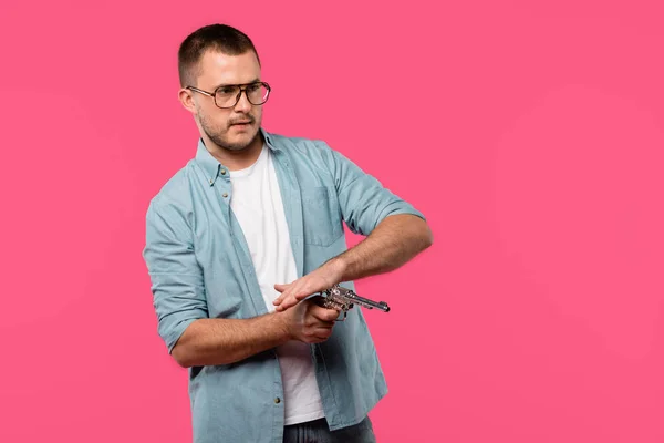 Молодий чоловік в окулярах перезаряджає револьвер і дивиться ізольовано на рожевий — стокове фото