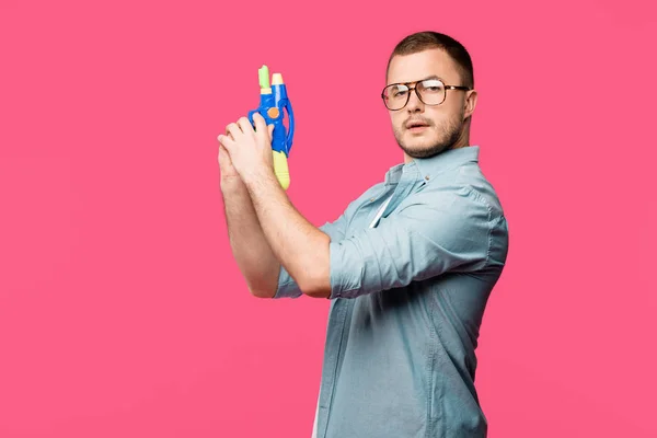 Молодий чоловік в окулярах тримає іграшковий пістолет і дивиться на камеру ізольовано на рожевому — стокове фото