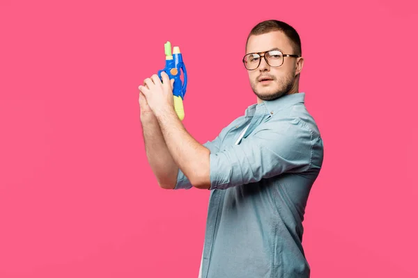 Joven en gafas con pistola de juguete y mirando a la cámara aislada en rosa - foto de stock