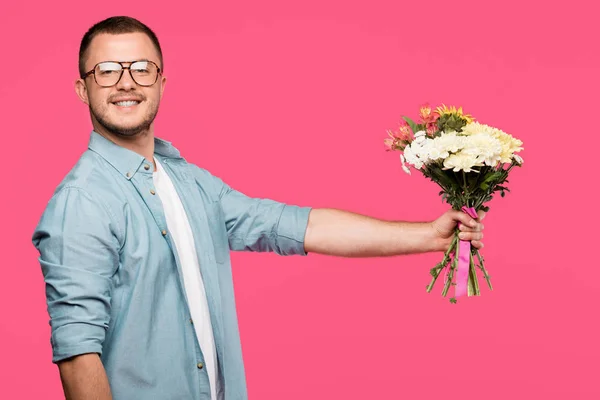 Jovem bonito segurando buquê de flores e sorrindo para a câmera isolada em rosa — Fotografia de Stock