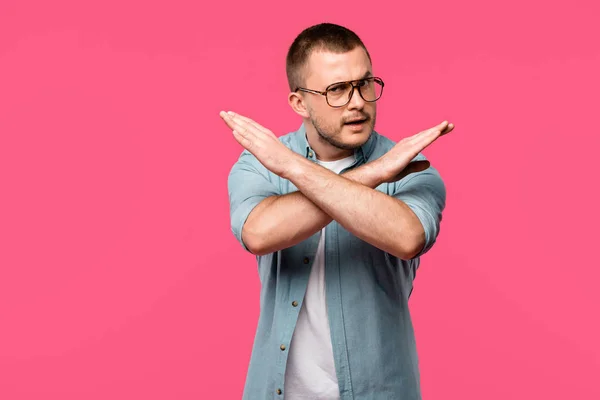 Junger Mann mit Brille gestikuliert Nein mit verschränkten Armen und blickt vereinzelt in die Kamera auf rosa — Stockfoto