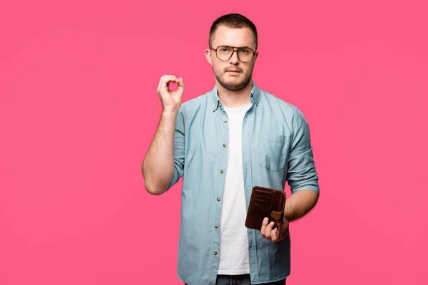 Молодий чоловік тримає порожній гаманець і показує знак нуля ізольовано на рожевому — стокове фото