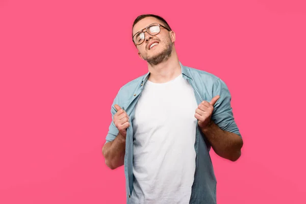 Cool young man in eyeglasses holding shirt and showing thumbs up isolated on pink — Stock Photo