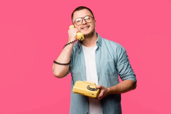 Smiling young man in eyeglasses talking by vintage telephone and looking at camera isolated on pink — Stock Photo