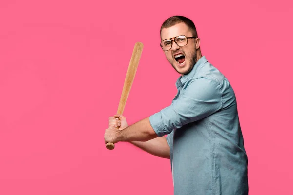 Aggressive young man in eyeglasses holding baseball bat and screaming at camera isolated on pink — Stock Photo