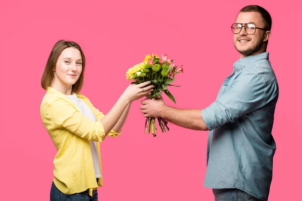 Seitenansicht eines glücklichen jungen Paares, das Blumenstrauß hält und isoliert auf rosa in die Kamera lächelt — Stockfoto